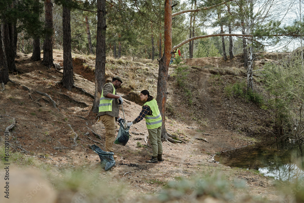 Fototapeta premium man and woman wearing safety vests collecting trash in forest near water, participating in environmental cleanup, working together to remove litter from natural area