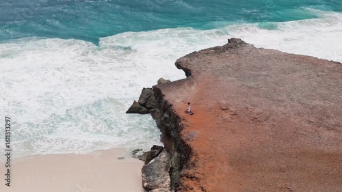 Toma aérea de dron de una pareja de jóvenes admirando y explorando el paisaje, sobre un acantilado rocoso y al fondo el mar azul con olas impactando en la costa de Aruba. Épico e inspirador.
