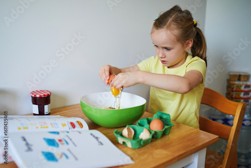 Adorable preschooler girl cooking a dish on herself. Little child consulting with recipe book and breaking an egg into a bowl.