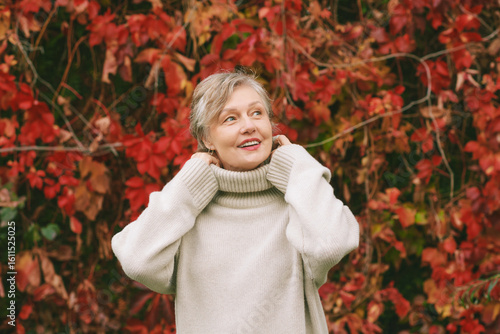Autumn portrait of happy mature woman wearing beige pullover or sweater, posing outdoors with red ivy red leaves background