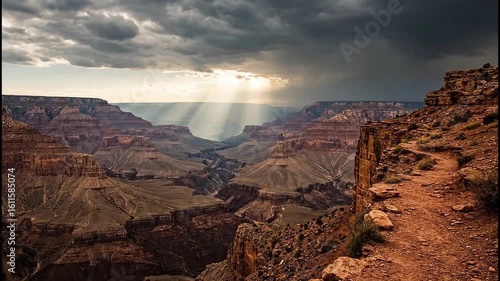 Dramatic sunbeams break through a stormy sky over the vast Grand Canyon, viewed from a cliffside trail.