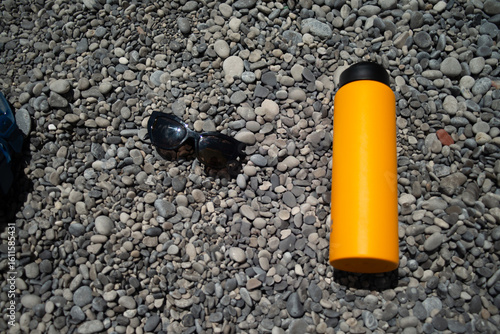 Εκτύπωση καμβά A refreshing summer scene featuring stylish sunglasses and a sleek water flask resting on smooth, sunlit pebbles at the edge of a serene beach