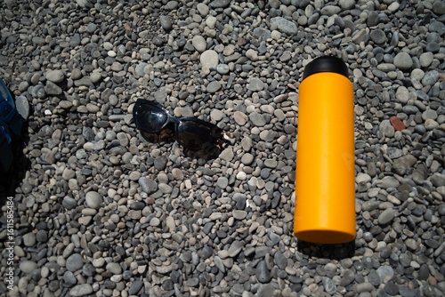 Bild auf Leinwand A refreshing summer scene featuring stylish sunglasses and a sleek water flask resting on smooth, sunlit pebbles at the edge of a serene beach