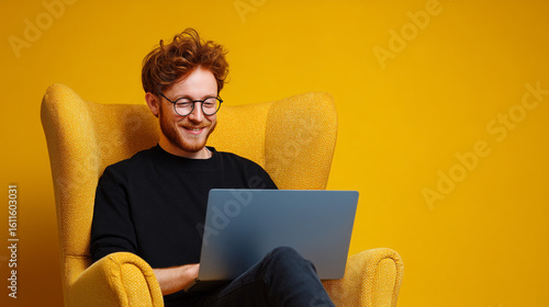 Man with glasses smiling while using laptop in yellow armchair against yellow background indoors