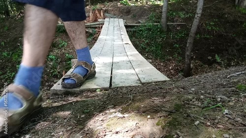 A man in sandals walks across a narrow wooden footbridge made of rough planks in a quiet forest. The simple structure blends with the natural surroundings, evoking a sense of solitude and nature.