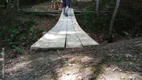 Man wearing sandals walks through a forest over a simple wooden plank bridge. Surrounded by trees and nature, the scene captures peaceful movement in the wild on a handmade footpath.