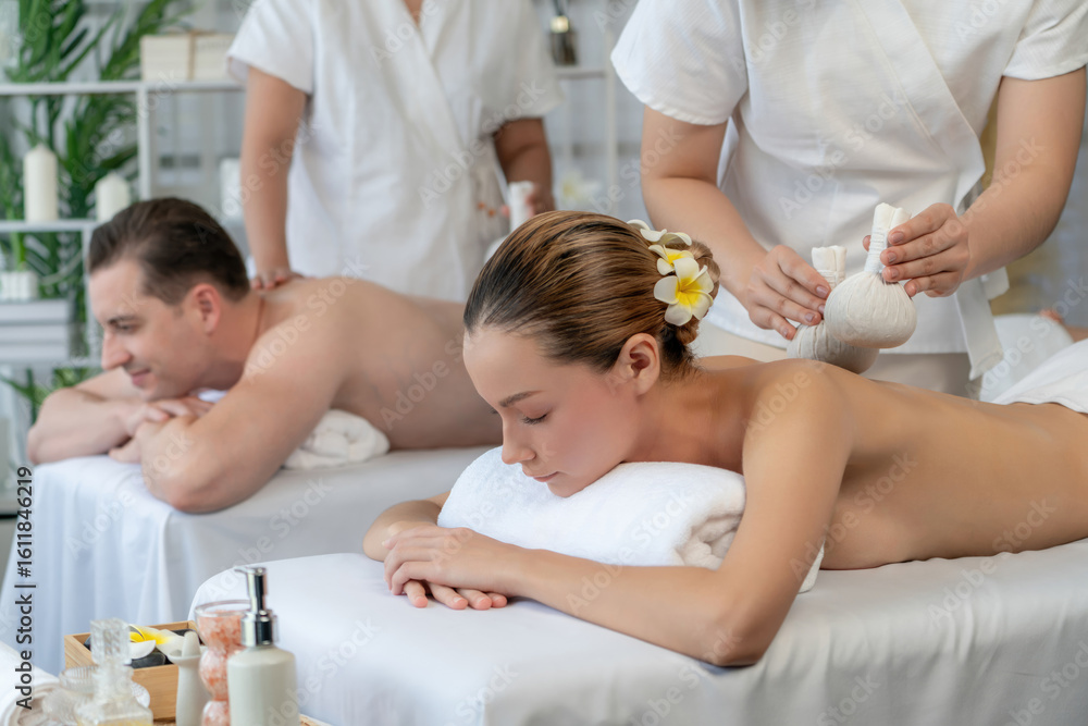 Obraz premium Hot herbal ball spa massage body treatment, masseur gently compresses herb bag on couple customer body. Tranquil and serenity of aromatherapy recreation in day lighting ambient at spa salon. Quiescent