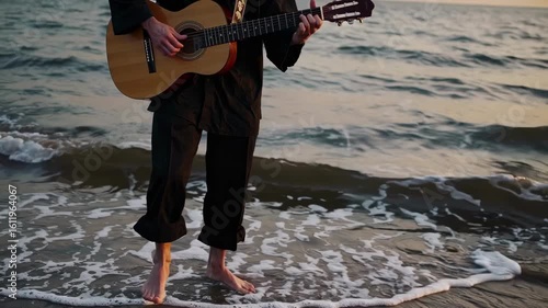 A man plays guitar by the sea at sunset, captured in a medium shot. The video conveys a serene, artistic vibe with natural lighting and a tranquil setting.