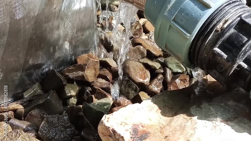 Water flows from a pipe onto rocks, creating natural splashes and gentle sound imagery. The scene blends industrial and natural elements, symbolizing drainage, irrigation, or nature reclaiming space.