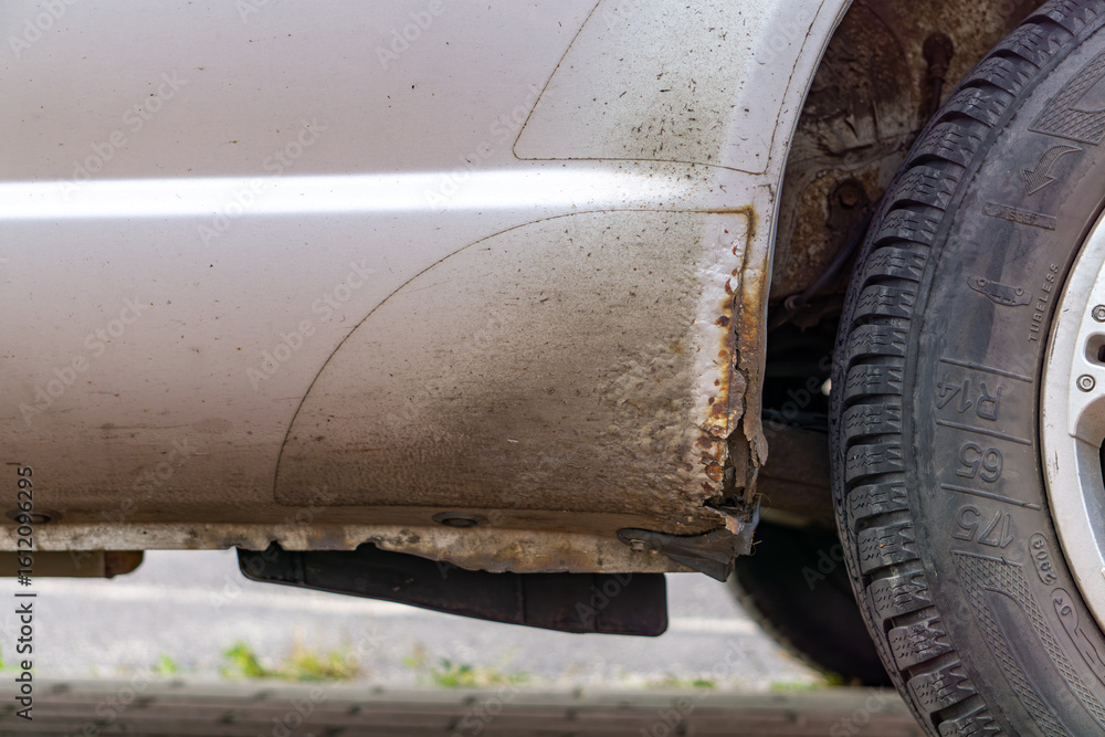 Visible rust and damage on the rocker panel of a silver passenger car. Close-up of corrosion near the rear wheel. Visible rust and damage on the rocker panel of a silver passenger car. Close-up of corrosion near the rear wheel.