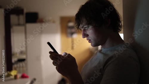 Man leaning against wall stares into smartphone with face glowing purple from screen light in dim room portraying digital immersion late night scrolling and emotional disconnect