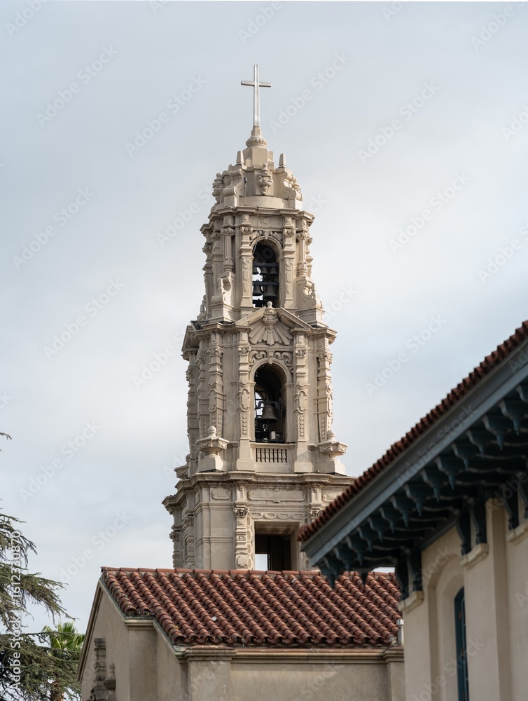 Fototapeta premium Ornate bell tower with a cross rises above red-tiled rooftops under a cloudy sky, showcasing classic Spanish-style architecture and historical charm.