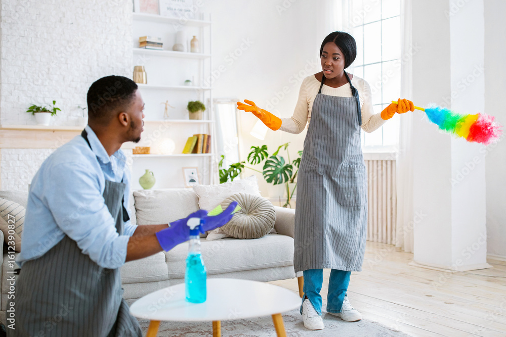 Fototapeta premium Quarrel over household chores. Angry black man and woman arguing while cleaning up their home, copy space. Millennial African American couple fighting over domestic duties