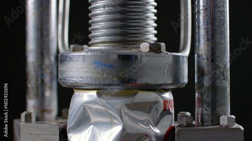 Close up of a metal can being crushed by a hydraulic press in a dark environment with metal supports
