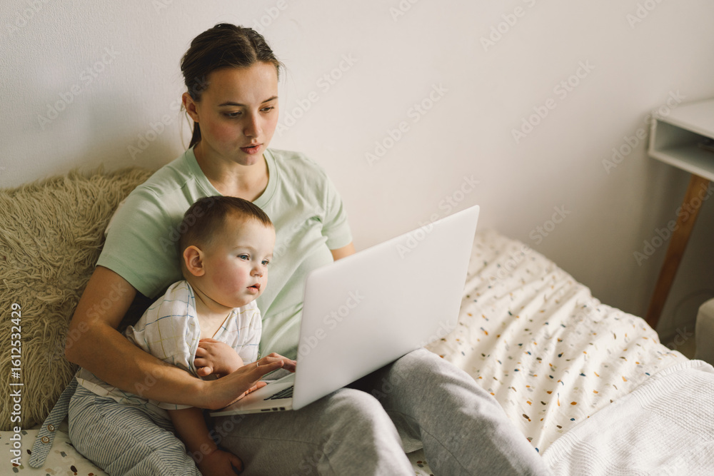 Portrait of happy young mother with little baby using laptop at home. Together, smiling mom browsing internet on computer while spending time with her little son. Portrait of happy young mother with little baby using laptop at home. Together, smiling mom browsing internet on computer while spending time with her little son.