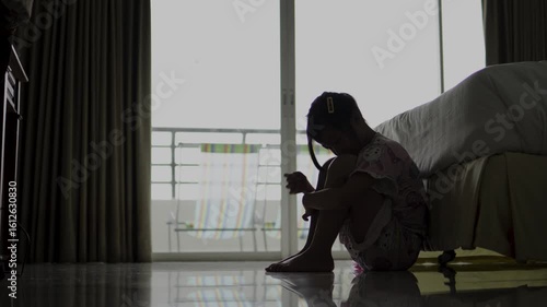 Young girl sitting on floor, hugging her knees in dimly lit room, reflecting on sadness and introspection as soft light from window enhances atmosphere.
