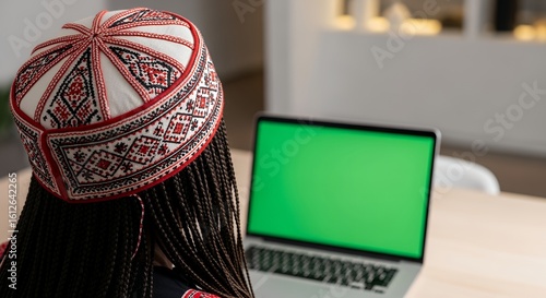 A woman wearing a traditional embroidered hat sits at a table using a laptop with a green screen.
