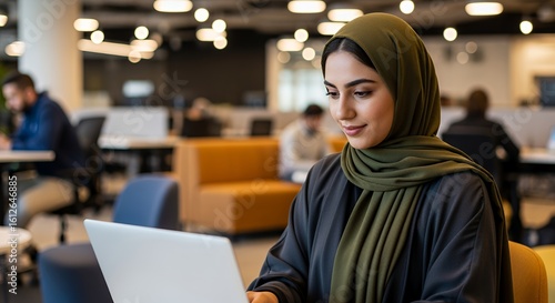 A young woman wearing a hijab works on her laptop in a modern, open-plan space.
