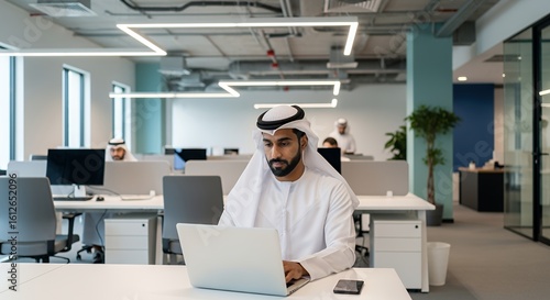 A young man in traditional Middle Eastern attire works diligently on his laptop in a modern, open-plan setting.