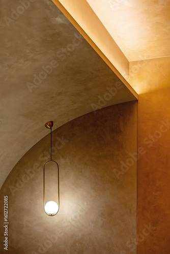A modern gold pendant light hangs against a curved, textured plaster wall with soft, warm cove lighting. Elegant and minimalist architectural detail.