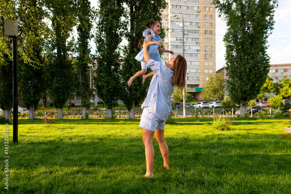 Fototapeta premium Mother in casual stripes joyfully hoists her giggling daughter in a blue dress into the air amid sunlit grass, trees, and apartment buildings on a warm summer evening