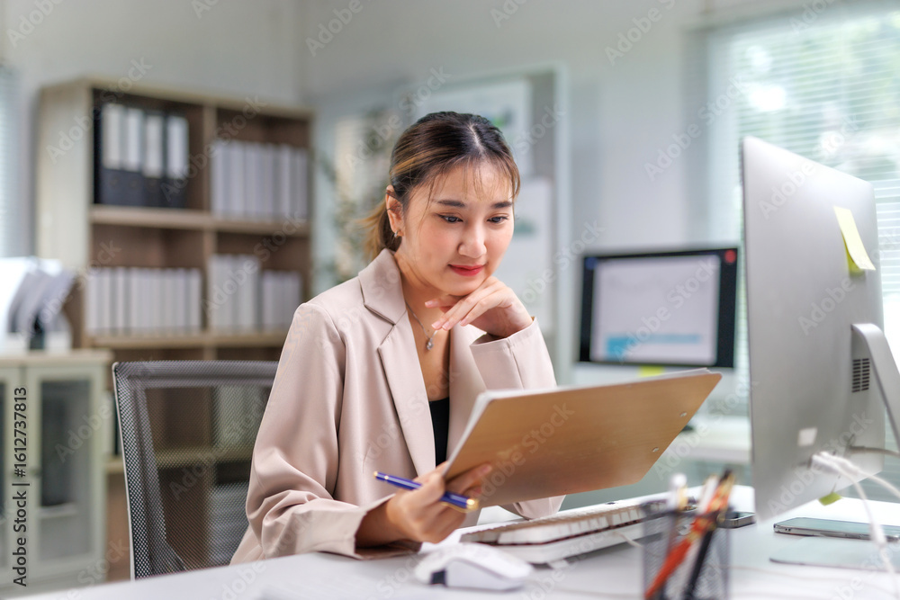 Young professional woman reviewing paperwork and working on a computer, maintaining focus in a bright, organized workspace that reflects modern design and efficiency Young professional woman reviewing paperwork and working on a computer, maintaining focus in a bright, organized workspace that reflects modern design and efficiency
