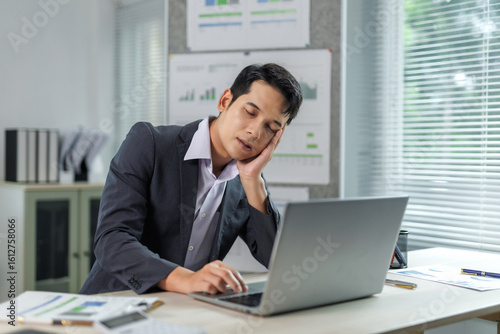 Obraz na plátně Young manager dozing off at workplace with hand on cheek, struggling to stay awa