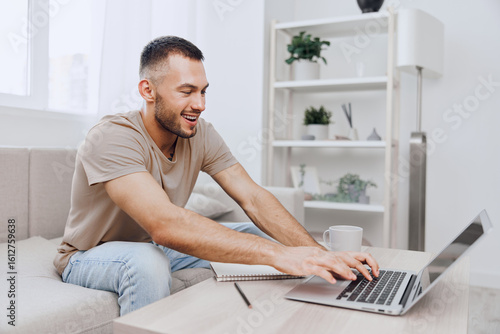 Free Download Smiling man engaged in remote work from home, gracefully typing on laptop, showcasing modern lifestyle and comfort in a cozy, bright living space. - GFXLoad