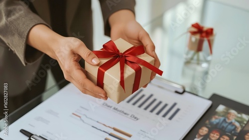 Woman hands holding red ribbon gift box, presenting surprise present, business office celebration, holiday event, blurred documents and charts background, warm natural light atmosphere