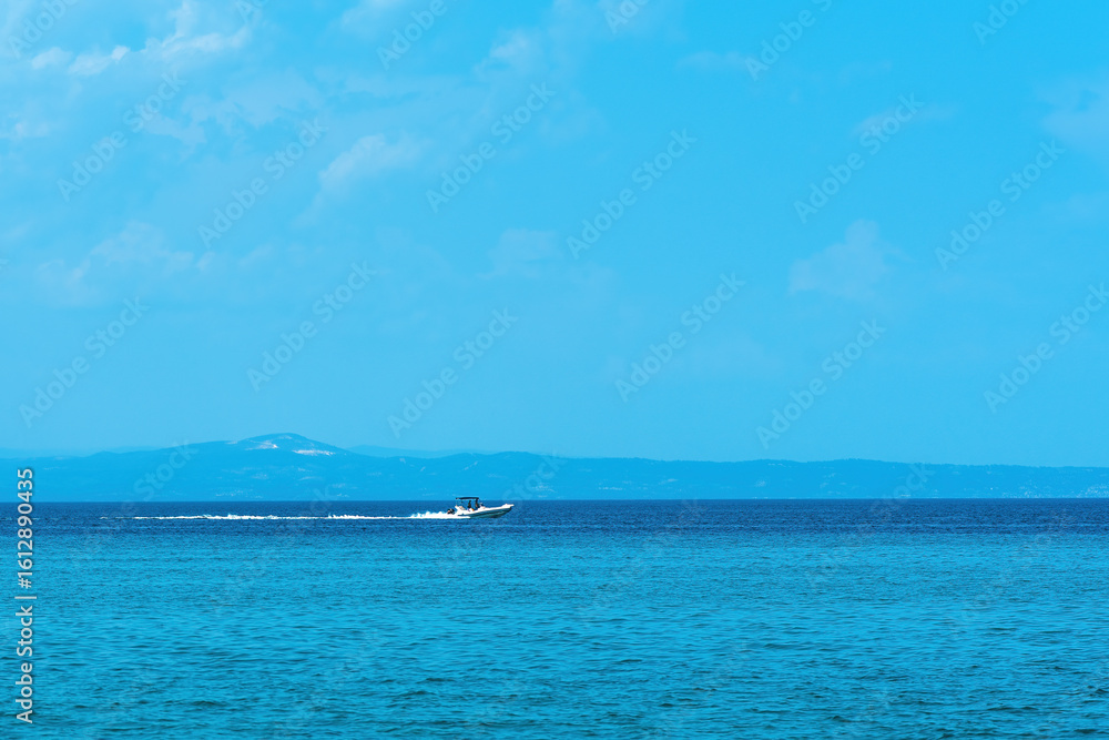 Fototapeta premium A small motorboat speeds across the vibrant blue sea, leaving a white trail in its wake. In the distance, faint outlines of hilly terrain stretch across the horizon