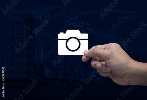 Fototapeta Naklejka Na Ścianę i Meble -  Hand holding camera flat icon in fingers over world map, modern city tower and skyscraper, Business camera service shop online concept, Elements of this image furnished by NASA