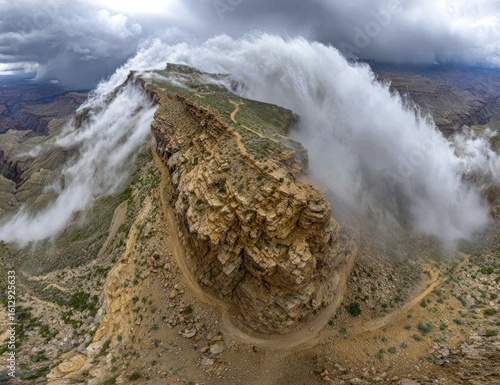 Dramatic canyon vista with low-lying fog cascading over a rugged, rocky ridgeline. A winding trail hugs the edge, offering views into the vast chasm under a stormy sky