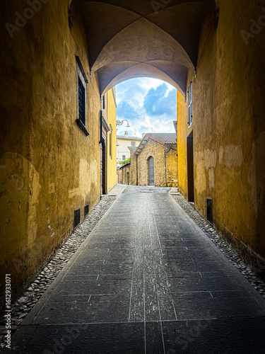 A narrow, historic alleyway in Spoleto, Italy, featuring a vaulted archway, ancient stone walls, and a glimpse of a bright sky at the far end, evoking old-world charm.