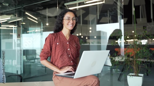 Happy young businesswoman working on laptop computer sitting at a desk in business office. Smiling female employee happily completing a task, chatting online with a colleague or browsing social media