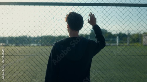Teenage boy stands facing sports fence lost in thought on sunny day