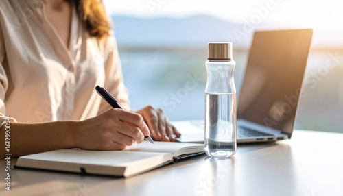Woman Writing in Notebook Near Laptop and Water Bottle