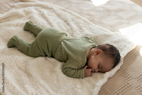 Infant girl wearing green onesie lying face down on white blanket over beige throw in bedroom