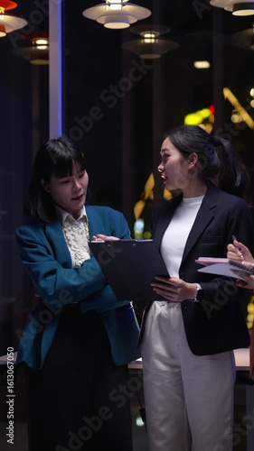 Professional Asian businesswomen collaborating during late evening office meeting, analyzing project details with serious focus, holding clipboards and notebooks near city window