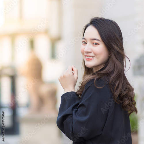 Square 1:1 portrait of beautiful Asian woman with long hair in black robe, smiling and walking while looking at camera outdoors in modern city background.