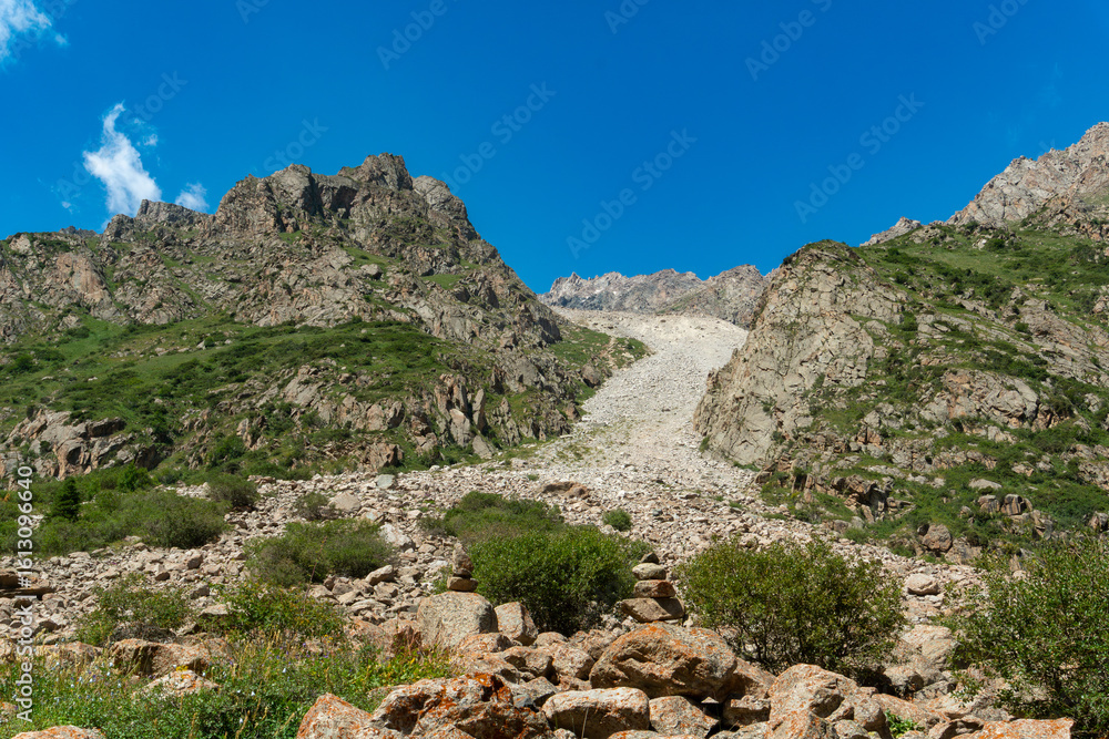 Naklejka premium Steep rocky mountains with a prominent scree slope, scattered boulders, green shrubs, clear blue sky, perfect for mountaineering, hiking, trekking, outdoor adventure, and nature exploration
