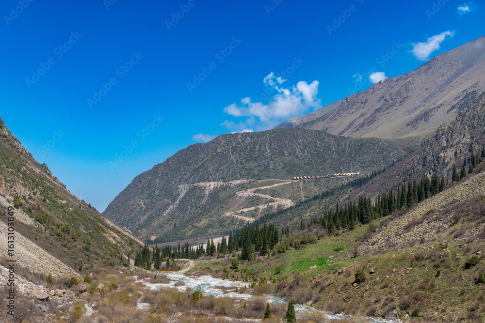 Fototapeta premium Ala-Archa Park, Mountainous terrain featuring a winding path, flowing river, pine trees, steep slopes, inviting setting for trekking, hiking, mountaineering, adventure, and nature exploration