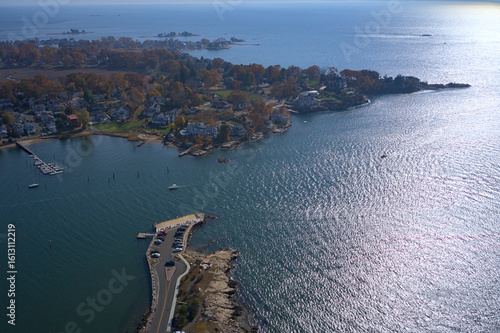 Aerial view of the sunlit water meeting the rocky coastline and autumn-tinged trees, with a pier stretching into the calm bay, Branford, Connecticut, United States.
