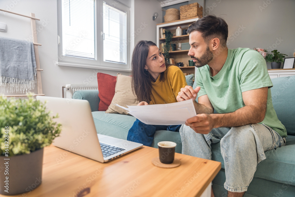 Fototapeta premium Doing Accounting at Home: Happy Couple Using Laptop Computer, Sitting at desk in Apartment. Young Family Filling Tax Forms, Mortgage Documents, Bills, Checks, Balances, Invoices are in Order