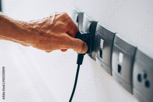Hand plugging a black electrical plug into a wall socket. Several sockets are aligned on a white wall in a modern interior.