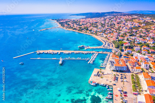 Drone image of Novalja on Pag island showing the coastal town layout, marina with anchored boats, clear Adriatic sea, waterfront buildings and sunlit Mediterranean surroundings