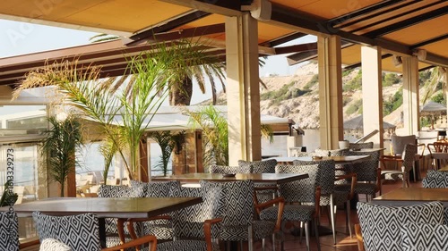 Wallpaper Mural Terrace of a coastal restaurant in Varkiza, Greece, with black and white patterned furniture, tropical plants, stone shore, calm sea, and bright sky. No people, peaceful summer atmosphere Torontodigital.ca
