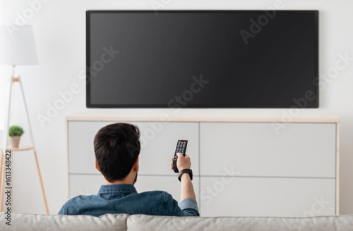 Photos Man watching television, turning on plasma flatscreen TV-set, pointing remote control at empty TV screen on white wall