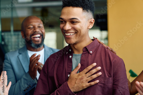 Grateful young male professional smiling while being applauded by colleagues in office