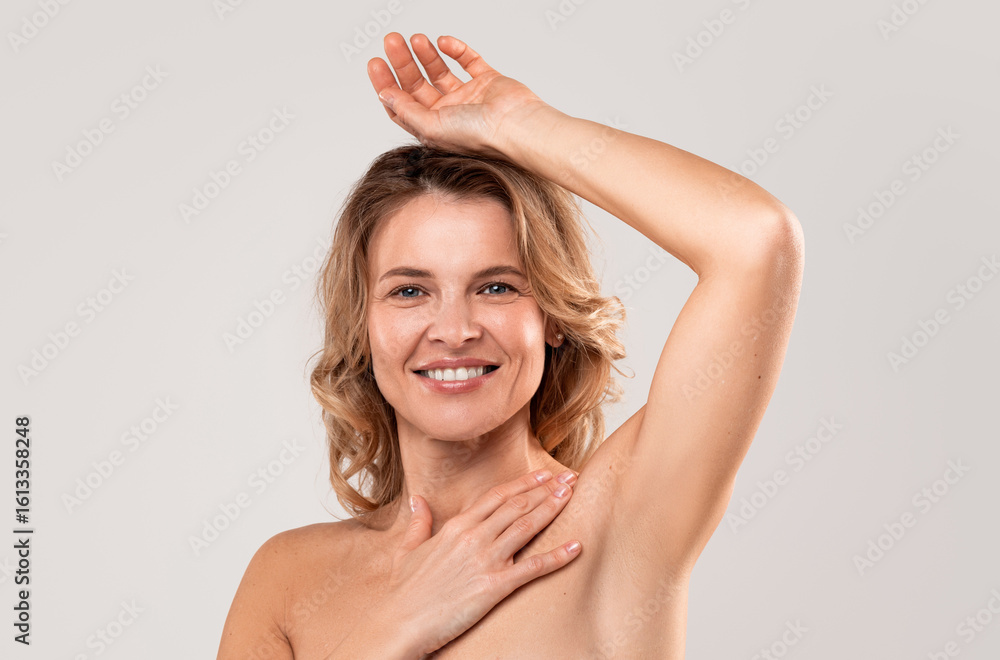 Obraz premium Portrait of beautiful middle aged lady showing her smooth hairless armpit at camera, attractive mature woman raising hand up and smiling, enjoying hair removal result, posing on light grey background