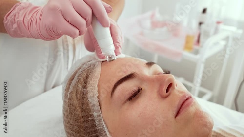 Patient woman receiving hydra facial treatment using advanced device at aesthetic center. Beautician focusing on forehead area with specialized equipment for skin hydration